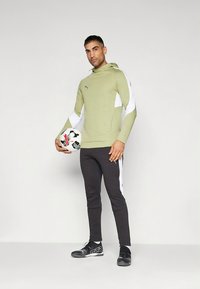 Sudadera con capucha verde y blanca con tela texturizada, combinada con pantalones negros ajustados y zapatos deportivos negros, sosteniendo un balón de fútbol.