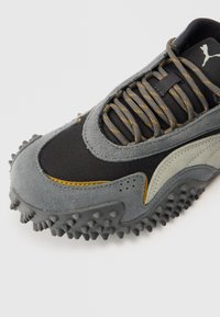 Grijze en zwarte sneaker met gestructureerd suède en mesh materialen, voorzien van gele accenten en een geribbelde rubberen zool, ontworpen voor grip en duurzaamheid.