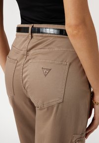 Ljusbruna byxor med en slät textur, med bakfickor och en trikornig logolapp. Ett svart strukturerat bälte förbättrar designen.