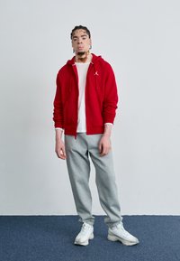 Haut à capuche zippé rouge avec poches avant, T-shirt blanc en dessous, pantalons de survêtement gris et baskets blanches ; tenue décontractée et athlétique.