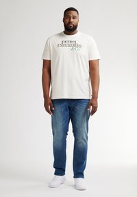 Camiseta de algodón de color beige claro con "PETROL INDUSTRIES & CO" impreso en verde y negro. Combinada con jeans azules desgastados y zapatillas blancas.