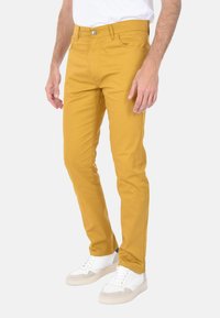 Pantaloni slim-fit gialli in cotone con cinque tasche, dotati di chiusura con bottone metallico e zip, abbinati a sneakers bianche.