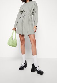 Vestito grigio a maglia con maniche lunghe e cinta, abbinato a una borsa intrecciata verde, calzini bianchi e scarpe platform nere.