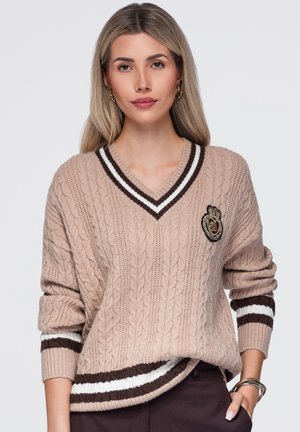 Une femme porte un pull beige à col V et à torsades, orné de rayures marron et blanches et d’un écusson, sur un fond clair uni.