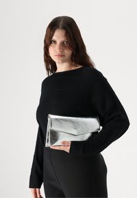 Geantă de mână metalică argintie, cu un design pliabil, ținută într-o mână. Geanta are o textură elegantă și accente minimaliste.