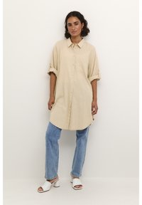Robe en lin beige à boutons sur le devant, avec des manches retroussées et un ourlet arrondi. Associée à un jean droit bleu clair et des sandales blanches à talons.