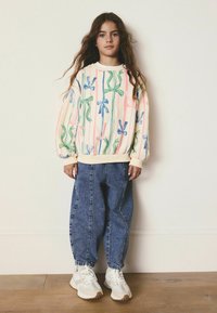 Sweatshirt mit einem Pastell-Hintergrund, der farbenfrohe Schleifen zeigt, kombiniert mit lockeren blauen Jeans und weißen Sneakers mit beigen Akzenten.