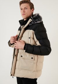 Giubbotto imbottito con parte superiore nera e parte inferiore beige, dotato di cappuccio in pelliccia sintetica, tasche frontali e bottoni a pressione. Tessuto testurizzato, design caldo.