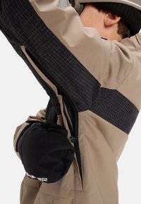 Dieťa v béžovej a čiernej bunde so bočným zipsom, čierny rukavice vložené do ventilačného otvoru podpažia a čierna pletená čiapka.