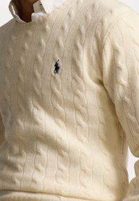 Krämfärgad kabelstickad tröja i mjukt material med V-ringning och en liten svart logotypaccents på bröstet.