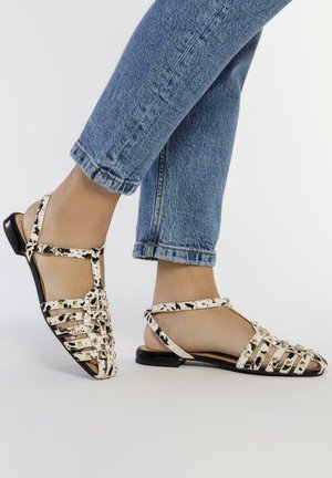 Sandalias blancas y negras con un patrón y correas entrecruzadas, tacón plano y punta abierta, combinadas con jeans azules remangados.