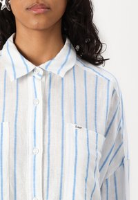 Balta krekls ar gaiši ziliem svītrām, ar apkakli, divām priekšējām kabatām un mazu "Lee" logo uz kabatas.