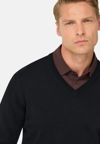 Pull noir en V en maille fine, porté sur une chemise à col marron. Vue rapprochée mettant l'accent sur le décolleté et la texture du tissu.