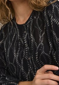 Femme portant un chemisier noir à boutons avec des motifs abstraits blancs et des cheveux blonds bouclés, la main posée sur son bras.