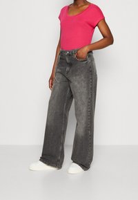 Grå brede jeans med falmet denimtekstur, parret med en lys pink top med scoop-udskæring og hvide snørresko, stående foran en ensfarvet baggrund.