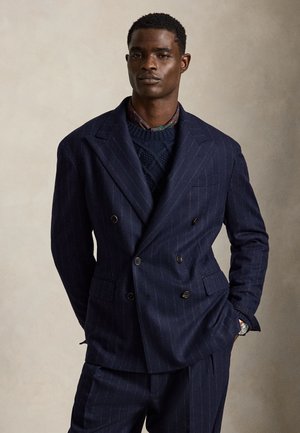 Homme portant un costume croisé bleu marine à fines rayures, un pull en maille bleu marine et un col de chemise à motif, debout les mains dans les poches.