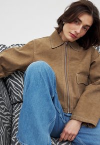 Giacca in suede marrone con chiusura a zip, ampio colletto e dettagli in cucitura bianca. Abbinata a jeans in denim a gamba larga di colore blu chiaro.