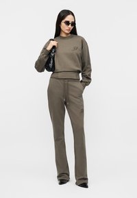 Γυναίκα που φοράει φούτερ σε ελιά πράσινο και παντελόνι που ταιριάζει, με μαύρη τσάντα χειρός και γυαλιά ηλίου, σε απλό λευκό φόντο.