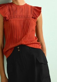 Top sin mangas de color óxido con textura acanalada y detalles de encaje, combinado con pantalones negros de cintura alta con bolsillos laterales y botones.
