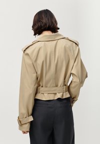 Beige cropped trenchcoat met schouderepauletten, riem in de taille en gerimpelde zoom. Soepele textuur en op maat gemaakte pasvorm. Zwarte broek is zichtbaar.