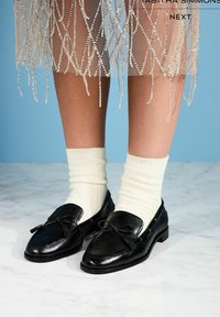 Schwarze Leder-Loafer mit einer Schleifendetail, getragen mit hellen, gerippten Socken. Die Schuhe haben eine abgerundete Spitze und einen niedrigen Absatz und sind auf einer Marmoroberfläche gezeigt.