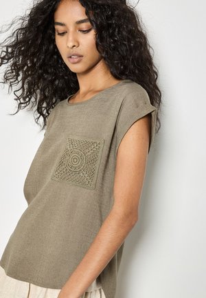 Donna con capelli lunghi e ricci che indossa una maglietta ampia a maniche corte color verde oliva con un disegno a tasca quadrata texturizzata, in piedi davanti a uno sfondo semplice.