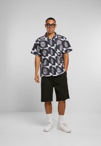 Kortermet svart-hvitt mønstrete skjorte med sirkulære design, paret med svarte shorts og hvite joggesko, står på en nøytral bakgrunn.