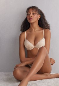 Soutien-gorge en dentelle beige avec un motif floral complexe, bretelles réglables et finition en tissu lisse. Modèle assis, jambes croisées sur une surface claire.