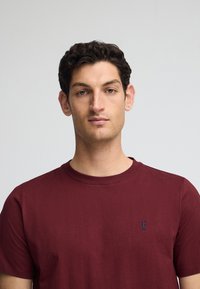 Bordeaux katoenen T-shirt met een ronde halslijn en korte mouwen; heeft een klein blauw geborduurd logo op de borst. Soepele textuur.