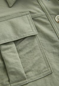 Camicia verde oliva con tessuto testurizzato, caratterizzata da una tasca sul petto, chiusura con bottoni e dettagli di cucitura visibili.
