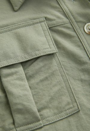 Olive green shirt with a textured fabric, featuring a chest pocket, button closure, and visible stitching details.