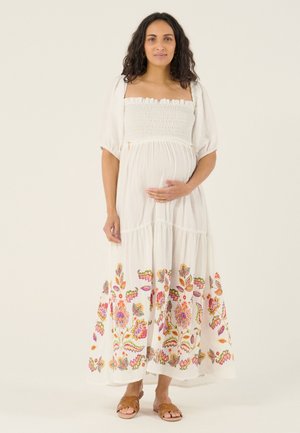 Femme enceinte portant une robe blanche avec une broderie florale le long de l’ourlet, tenant son ventre, debout devant un fond uni.