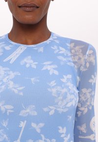 Top à manches longues bleu clair avec un motif floral, fabriqué en tissu transparent et texturé. Présente un col rond et des détails de couture visibles.