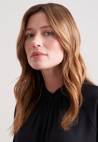 Blusa nera con colletto rotondo, caratterizzata da pieghe sottili e chiusura a bottoni. Capelli lunghi e ondulati di colore marrone incorniciano parzialmente il viso.