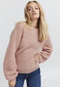 Maglione rosa lavorato a maglia con un motivo testurizzato, maniche ampie e scollo a girocollo. Abbinato a jeans in denim blu.