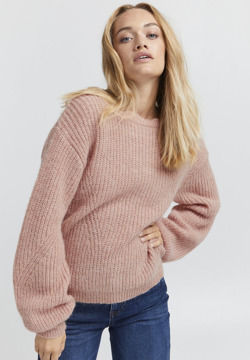 Rosa gestrickter Pullover mit strukturiertem Muster, weitem Schnitt und Rundhalsausschnitt. Kombiniert mit blauen Jeans.