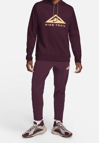 Sweat à capuche Nike Trail bordeaux avec un grand logo doré, assorti à un pantalon bordeaux. La tenue présente une coupe décontractée et un design athlétique.
