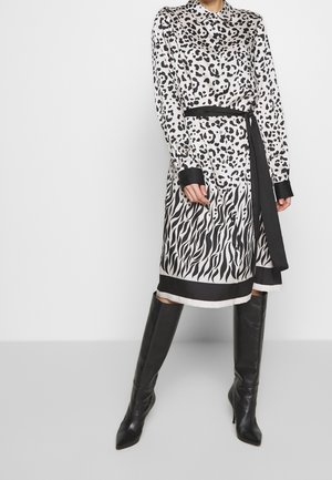 Frau trägt ein langärmliges, geknöpftes Kleid mit schwarz-weißem Tiermuster, schwarzen Gürtel und kniehohe schwarze Lederstiefel.
