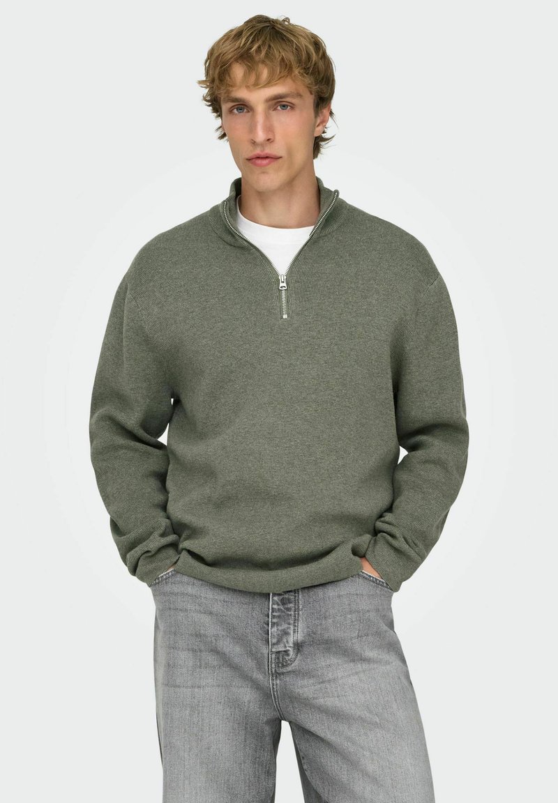 Groene trui met ritssluiting, voorzien van een gestructureerd breipatroon, geribbelde manchetten en een opstaande kraag, gedragen over een wit shirt en gecombineerd met grijze spijkerbroeken.