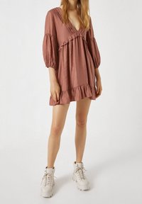 Kurzes pinkes Kleid mit Puffärmeln und Rüschendetails, das einen tiefen V-Ausschnitt und einen ausgestellten Saum hat. Kombiniert mit klobigen weißen Sneakern.