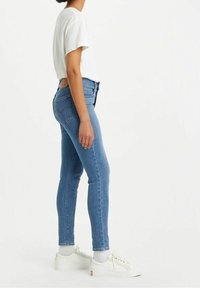 Jeans ajustados de color azul fabricados en denim, con una cintura alta, cinco bolsillos y un parche de piel en la parte posterior. Llevados con una camiseta blanca y zapatillas blancas.