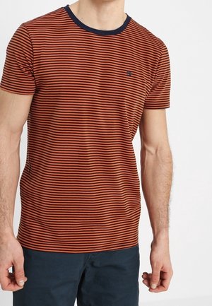 Mann trägt ein kurzärmeliges T-Shirt mit rost- und schwarzfarbenen Streifen, einem marineblauen Kragen und dunkelblauen Hosen, steht vor einem einfarbigen Hintergrund.