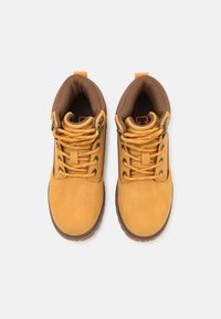 Botas de ante amarillas con acentos marrones, punteras redondas y un interior marrón con patrón. Incluyen cordones amarillos y ojales de metal.