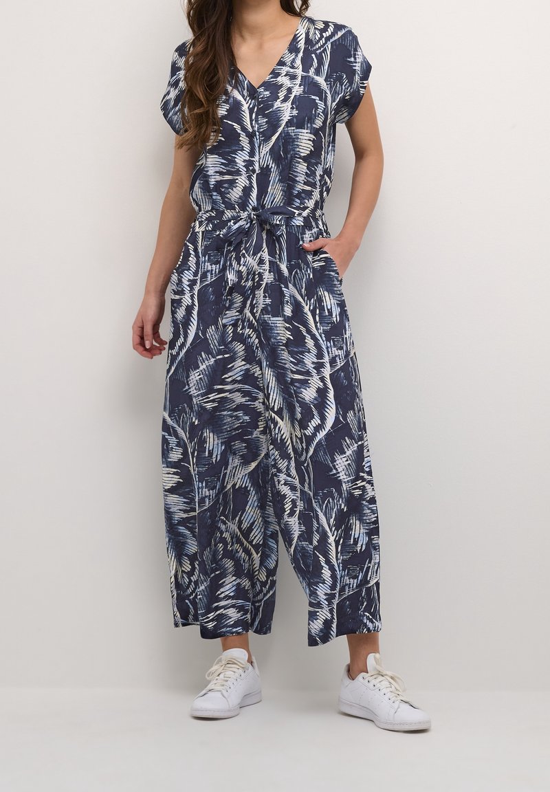 Femme vêtue d’une combinaison bleu marine et blanche à motif abstrait, manches courtes et baskets blanches, debout contre un mur uni.