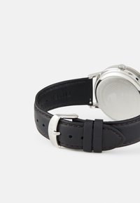 Correa de reloj de cuero negro con hebilla de metal plateado y detalles de costura. Caja de reloj redonda con una superficie lisa y reflectante.