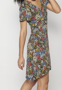 Femme portant une robe courte à manches courtes et longueur genou avec un motif floral coloré sur fond noir.