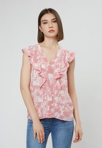 Rosa Bluse mit floralem Muster, Rüschensäumen und Spitzenbesatz, mit V-Ausschnitt und vertikalen Knopfdetails. Getragen mit blauen Jeans.