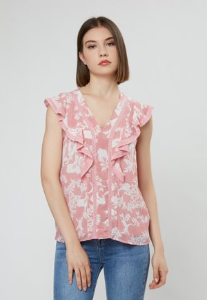 Roze bloemenblouse met gerimpelde mouwen en kantafwerking, met een V-hals en verticale knopen. Draagt met blauwe jeans.