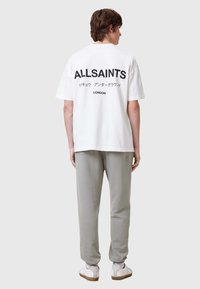 Vit bomull t-shirt med svart text "ALLSAINTS" och "LONDON," ihop med grå joggers och vita sneakers med en beige sula.