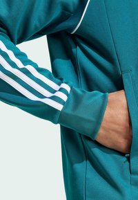 Teal mikina s bílým třípruhovým designem na rukávech, hladká textura a přední kapsa. Disponuje jemným leskem a sportovním střihem.